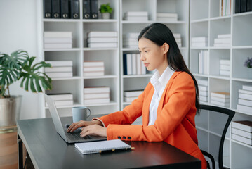  Beautiful Asian woman using laptop and tablet while sitting at her working place. Concentrated at work.