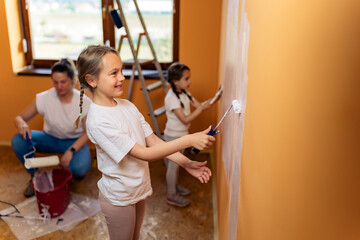 The older daughter is painting the wall and posing for the camera, the mother and younger daughter in the background