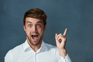 Portrait, wow and idea with a man pointing in studio on a blue background during a lightbulb moment. Face, surprise and eureka with an excited young male person looking shocked or amazed by good news