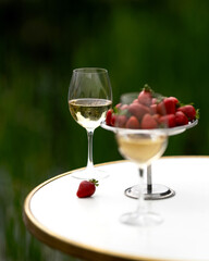 Two glasses of white wine and bowl of strawberries on table. Picnic. Ripe berries. Wine tasting. Vineyard. White table. Blurred green background. Side view. Copy space. Soft focus. 