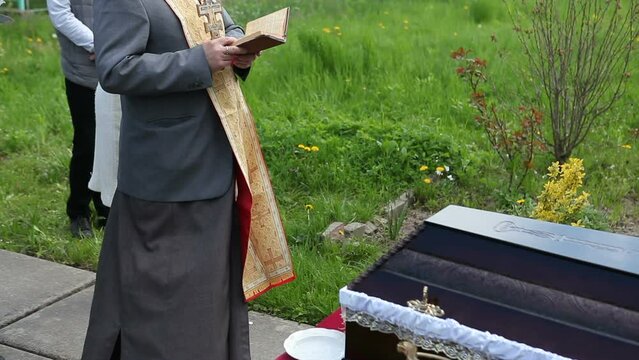 A closed coffin with a dead man stands in a cemetery. A priest with a cross in his hands buries the deceased, reads a prayer.