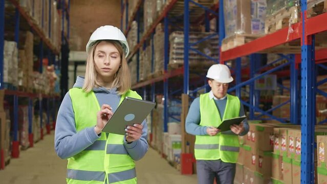 Happy caucasian woman working on digital tablet and shaking hands with asian coworker during meeting at factory storehouse. Two competent inspectors greeting each other among shelves with goods.