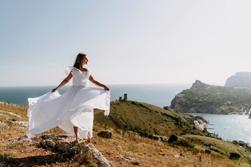 Naklejka na ściany i meble Happy woman in a white dress and hat stands on a rocky cliff above the sea, with the beautiful silhouette of hills in thick fog in the background.