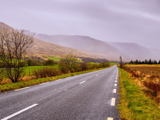 Small narrow asphalt road to mountains in low cloudy sky. Travel background. Calm Irish nature landscape. Transportation and tourism concept. West of Ireland.