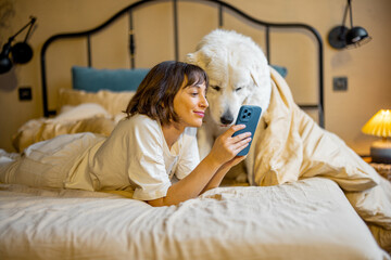 Young woman uses smart phone while lying with her cute adorable dog in bed at cozy bedroom in beige tones. Concept of leisure time with pets