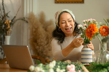 Cheerful senior woman having video call on laptop and arranging flowers in vase in living room....