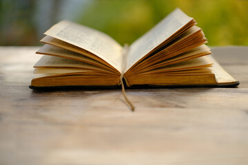 open paper book on old wooden table in garden, beautiful blurred natural landscape in background with green foliage, concept education, nostalgia, knowledge