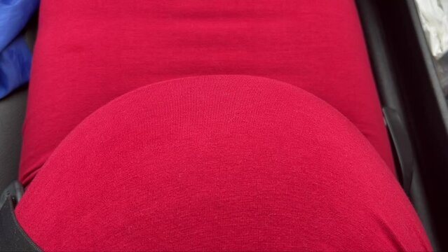 mom-to-be, wearing a red dress, seating in car with seat belt on and baby kicking and moving inside pregnant belly while riding in the car
