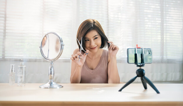 Asian woman show how to cut hair with scissors to fan following channel record video camera at home. Online influencer teach girl social media market live steam barber do it yourself minimal lifestyle