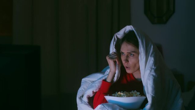 Portrait of pretty emotional woman watching scary movie horror thriller on TV streaming with scared faces holding bowl of popcorn sitting on the sofa at home alone covered with a white blanket. 