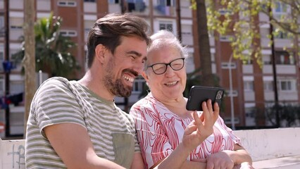 Adult grandson teaches his grandmother how to use smartphone. Sitting outdoors taking a video call together speaking with family.