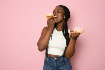 Hungry Afro woman bites tasty doughnut with bright sprinkles, has unhealthy nutrtion, cant imagine life without sweet dessert isolated on pink background. - obrazy, fototapety, plakaty