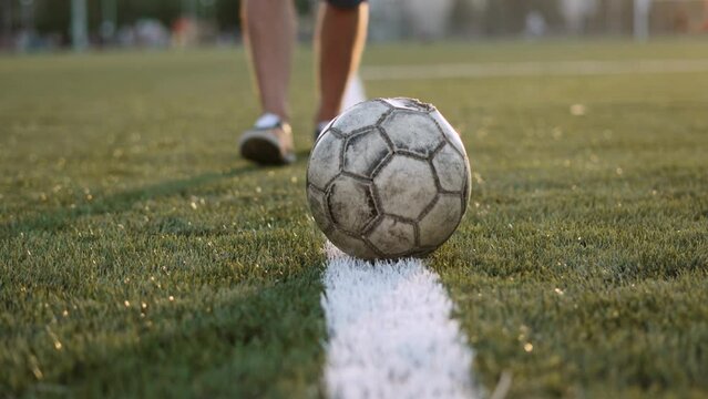 Unrecognizable football player passes old shabby soccer ball, close-up. Ball rolls along white line of penalty area of football field at sunset. Blurry man with hairy legs follows sports equipment.