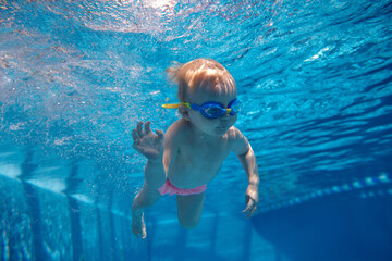 Cute, funny, little baby girl, toddler in goggles swimming underwater in swimming pool. Water...