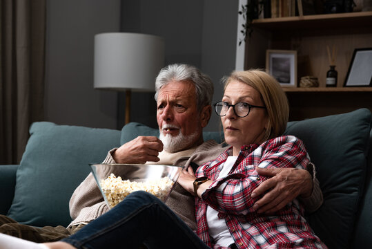 Mature married couple entertaining observing terrified TV program. Senior people watching horror movie at home with popcorn making scared faces. Older husband and wife watch television