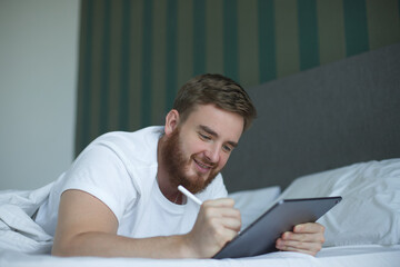 Young happy man use tablet laptop computer at home in bed, lay in bedroom, looking at screen of gadget. Concept of freelance work, distant job, online shopping or education