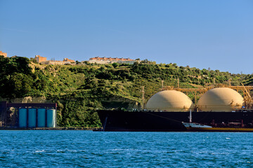 LNG tanker moored by industrial pier and green hill on sunny day