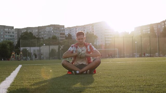 Portrait of upset soccer player sitting in lotus position on urban football pitch at sunset. Man sadly squeeze old, shabby ball with puncture in hands. Concept of bad quality equipment, end of game.