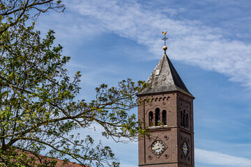 Bell tower of Our Lady Immaculate Conception church in neo-Romanesque style against blue sky, brick walls, clock and green foliage of a tree, spring sunny day in Susteren, South Limburg, Netherlands