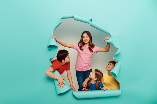 Smiling interracial kids in colorful clothes looking at friend while celebrating international children day behind hole in blue paper background