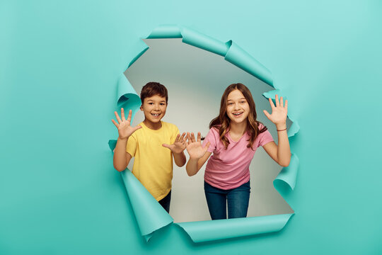 Smiling multiethnic kids in colorful t-shirts waving hands at camera while celebrating world protection children day near hole in blue paper background