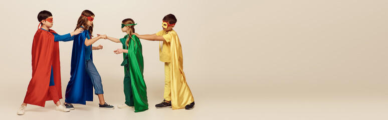 side view of multicultural kids in colorful superhero costumes with masks and cloaks fighting with each other on grey background in studio, International children's day concept, banner