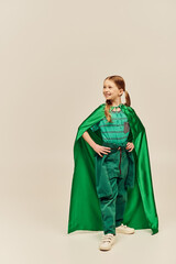 smiling girl in green superhero costume with cloak wearing pants and t-shirt and standing with hands on hips while celebrating International Children Day on grey background