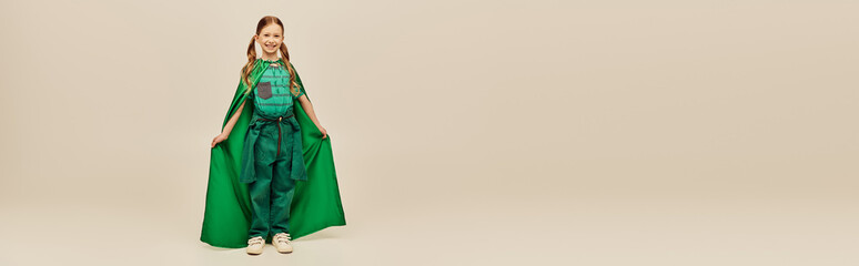 smiling girl in green superhero costume with cloak wearing pants and t-shirt and standing while celebrating Child protection day holiday on grey background, banner