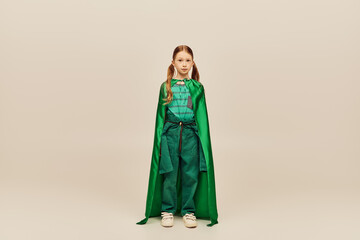 Redhead preteen girl in green superhero costume and cape looking at camera while standing on grey background during global child protection day celebration