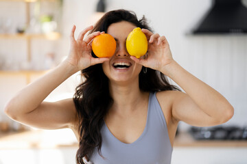 Portrait of beautiful smiling latin woman holding lemon and orange near face standing in modern kitchen. Healthy lifestyle concept