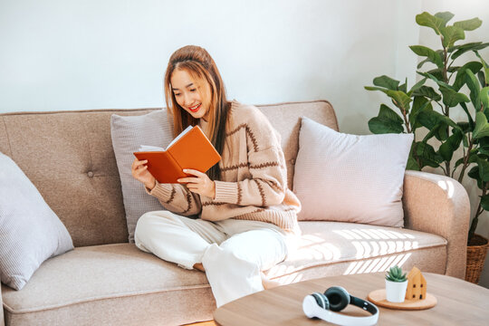 Pensive Relaxed Asian Woman Reading A Book At Home Relax And  Drinking Coffee On Sofa.