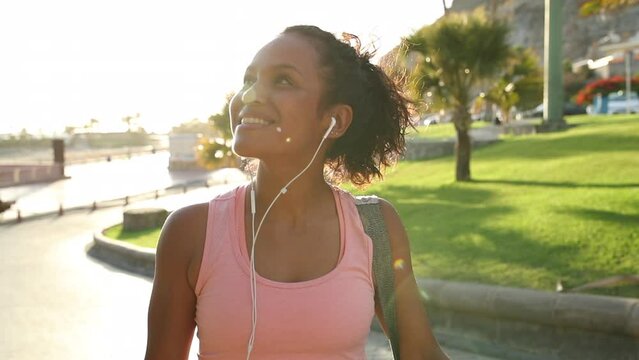 Smiling young African woman in sportswear listening to music on earphones while crossing the street carrying a yoga mat