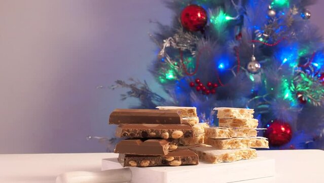 Sugar and Chocolate Turron with nuts. Traditional Spanish almond dessert on wooden table. Nougat candy on the background of a decorated Christmas tree with a flashing garland.