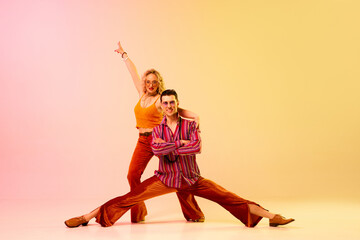 Dynamic image of stylish, talented, emotional couple, man and woman in vintage costumes dancing disco dance over gradient pink yellow background. Retro style, fashion, art, hobby, music, 70s concept