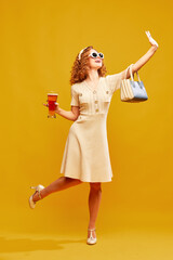 Young charming girl wearing stylish retro clothes with sunglasses holding glass of beer over yellow background. Alcohol festival
