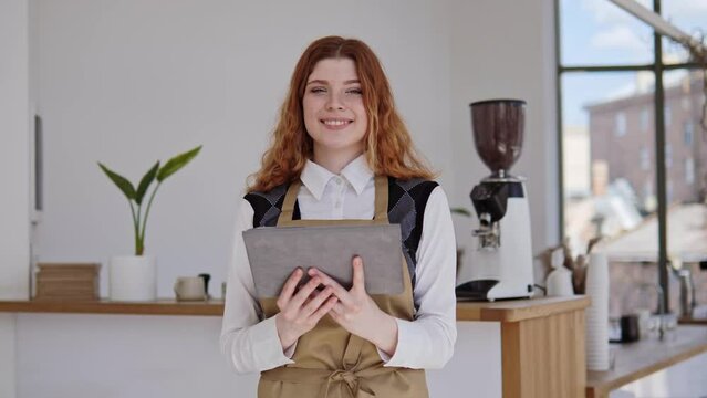 Barista Portrait, Beautiful Female Red Hair Salesperson, Small Business Owner Of Cafe Dressed In Apron And White T-shirt, Holding A Tablet Takes Orders For Her Coffee Shop, Looking To Camera Smiling.