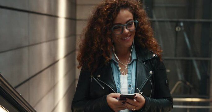 Stylish young woman smiling and listening to music on her cellphone while riding up a metro station escalator 