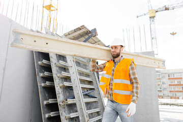 Builder man in hardhat and vest carrying timber on building site in his working day