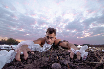 sports man with athletic figure wrapped in plastic on the ground at sunset, garbage problem,...