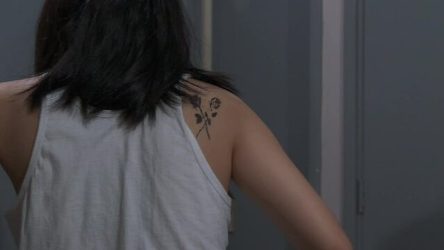 Girl with medium hair takes off black jacket to reveal red devil and flower tattoos, ties hair up