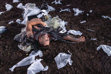 sports man with athletic figure wrapped in plastic on the ground at sunset, garbage problem, environmental disaster, land pollution
