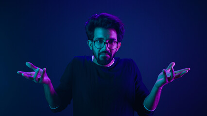 Studio dark blue neon background Indian man Arabian male model guy confused shrug shoulders unsure doubtful shrugging doubt uncertain don't know problem solution question guess puzzled bemused no clue