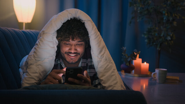 Smiling guy Indian man Arabian male at night evening home on couch under blanket cover with duvet looking mobile phone wow surprised amazed reading news gadget addict smartphone social media scrolling