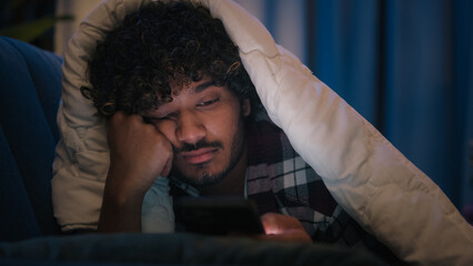 Sad sleepy guy Latino tired man upset Arabian sleeping napping boring Indian male at night evening dark home under blanket cover with duvet scrolling mobile phone addict smartphone bored social media