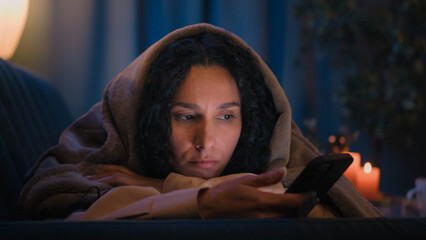 Hispanic sad sleepy girl Latino tired woman upset boring Indian lady bored Arabian female night evening dark home under blanket cover with duvet scrolling mobile phone addict smartphone social media