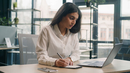 Multiracial serious businesswoman in office working with laptop writing notes Indian girl Arabian...