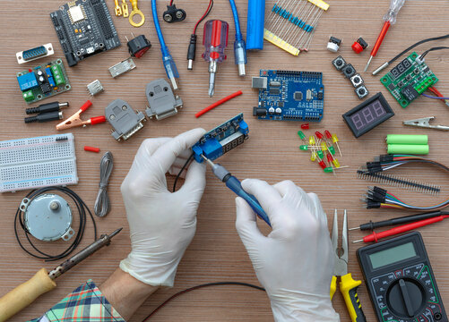 An engineer's hands are assembling a breadboard from an Arduino microcontroller, surrounded by electronics tuning tools. View of the electronic laboratory bench with peripherals and expansion boards.