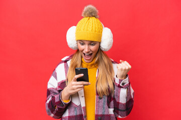 Young beautiful woman wearing winter muffs isolated on red background surprised and sending a...