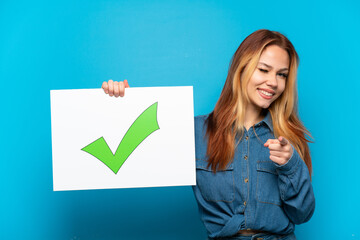 Teenager girl over isolated blue background holding a placard with text Green check mark icon and pointing to the front