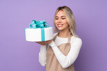 Young pastry chef holding a big cake over isolated purple background